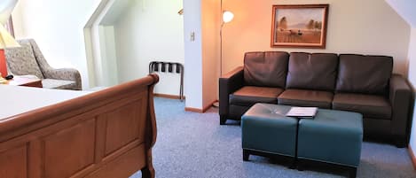 A room with a wooden bed, a brown leather sofa, a green ottoman, a framed picture on the wall, and a floor lamp.