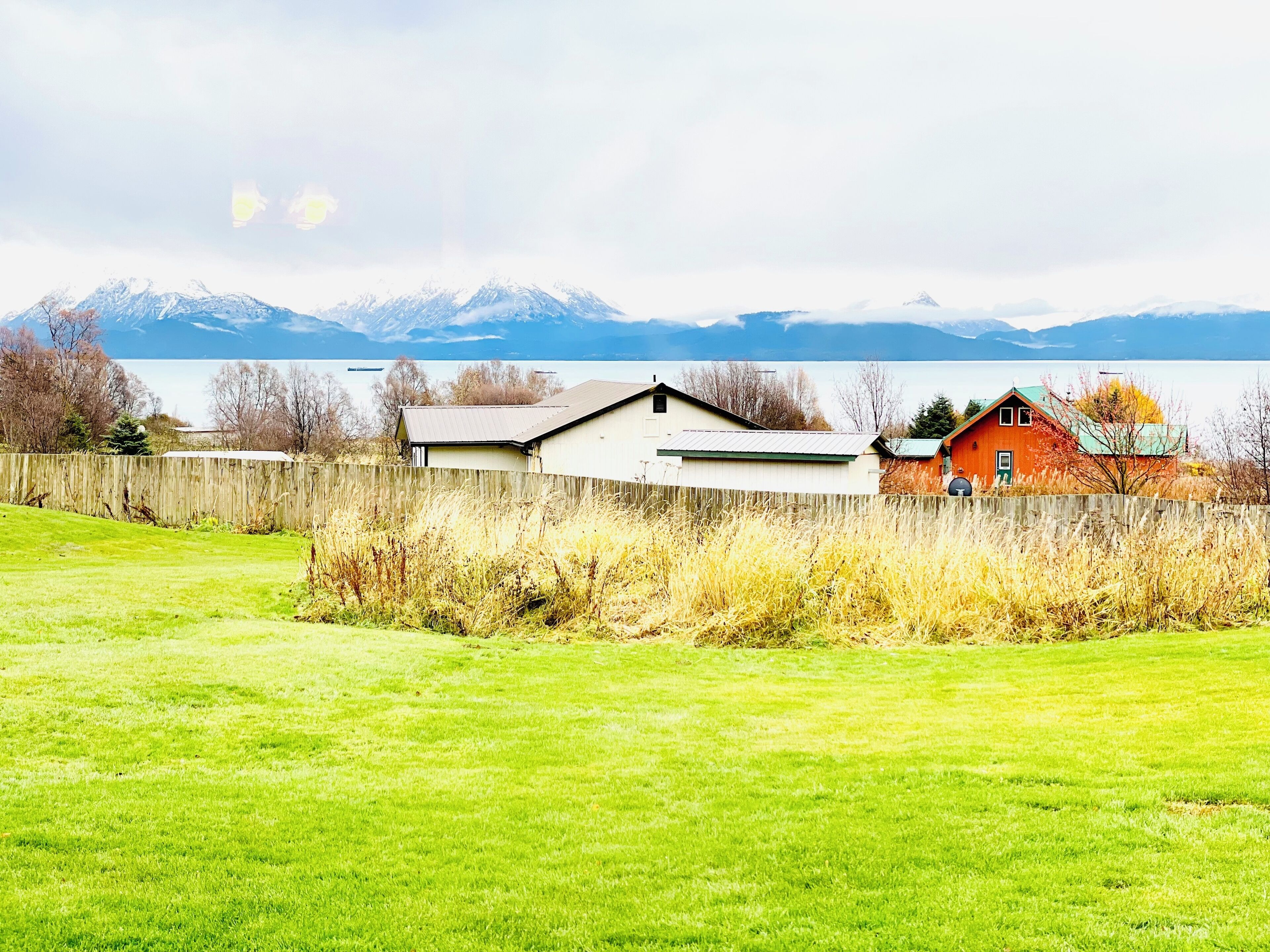 Ett lantligt landskap med ett trästaket, en gräsklädd åker och en rad hus, med en sjö och berg i bakgrunden.