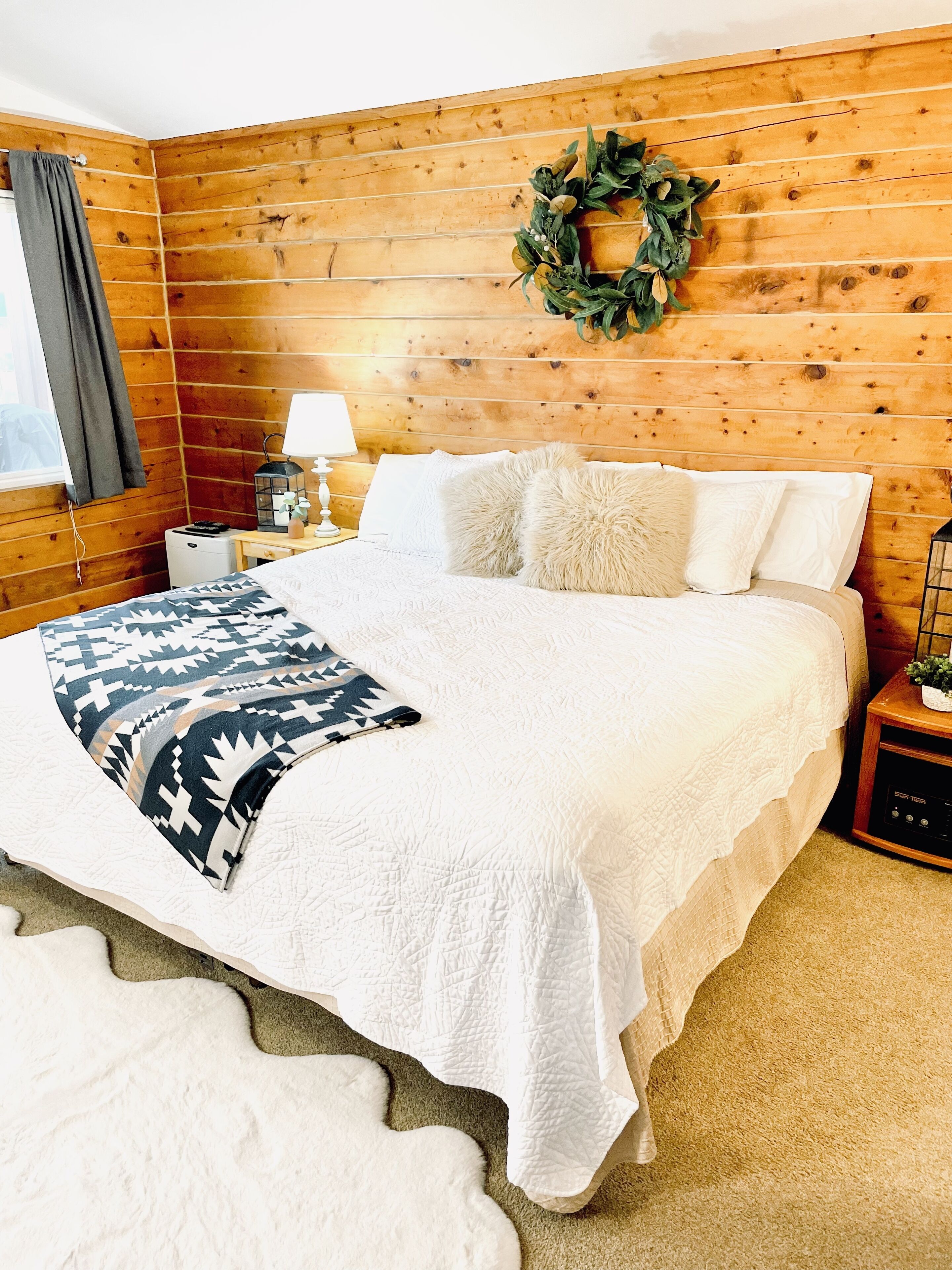 A bedroom with a wooden paneled wall, a bed with white bedding, a decorative wreath, and a nightstand with a lamp and a small plant.