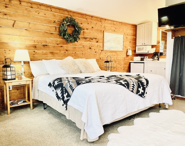 Una habitación acogedora con pared de madera, una cama con ropa de cama blanca, una mesita de noche con una lámpara y un televisor en una esquina.