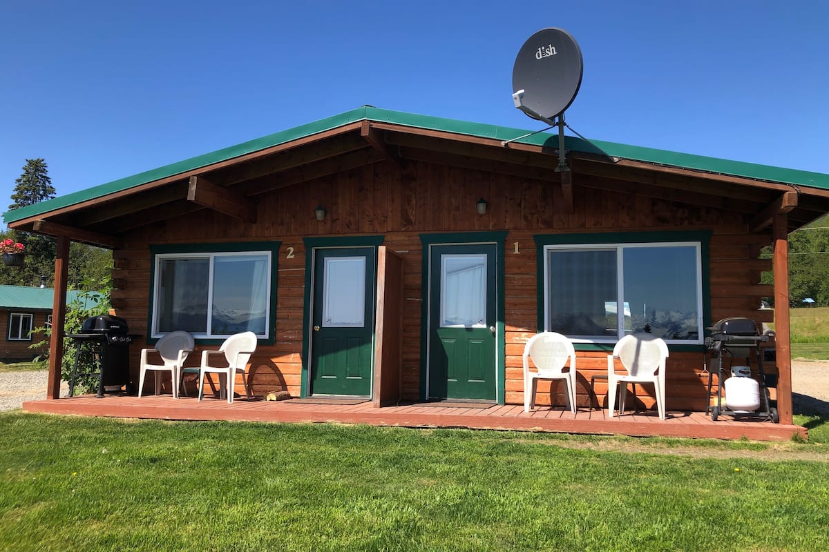 A wooden cabin with two doors, a satellite dish, and a patio with chairs and a grill.