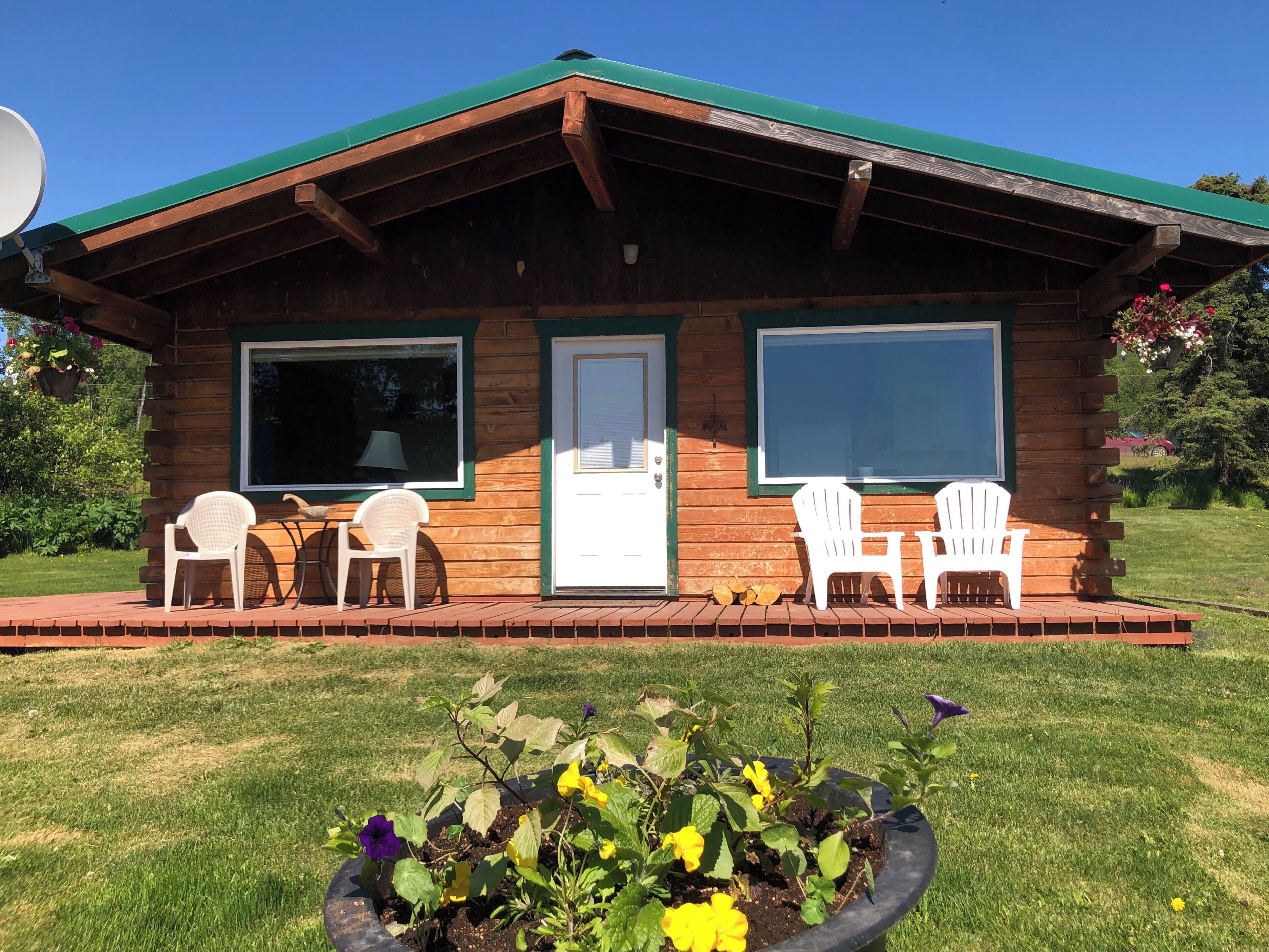 A wooden cabin with a green roof, two windows, and a porch with white plastic chairs.