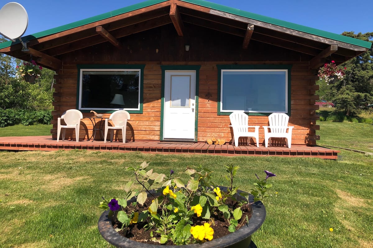 A wooden cabin with two windows, a door, and a porch with white chairs.