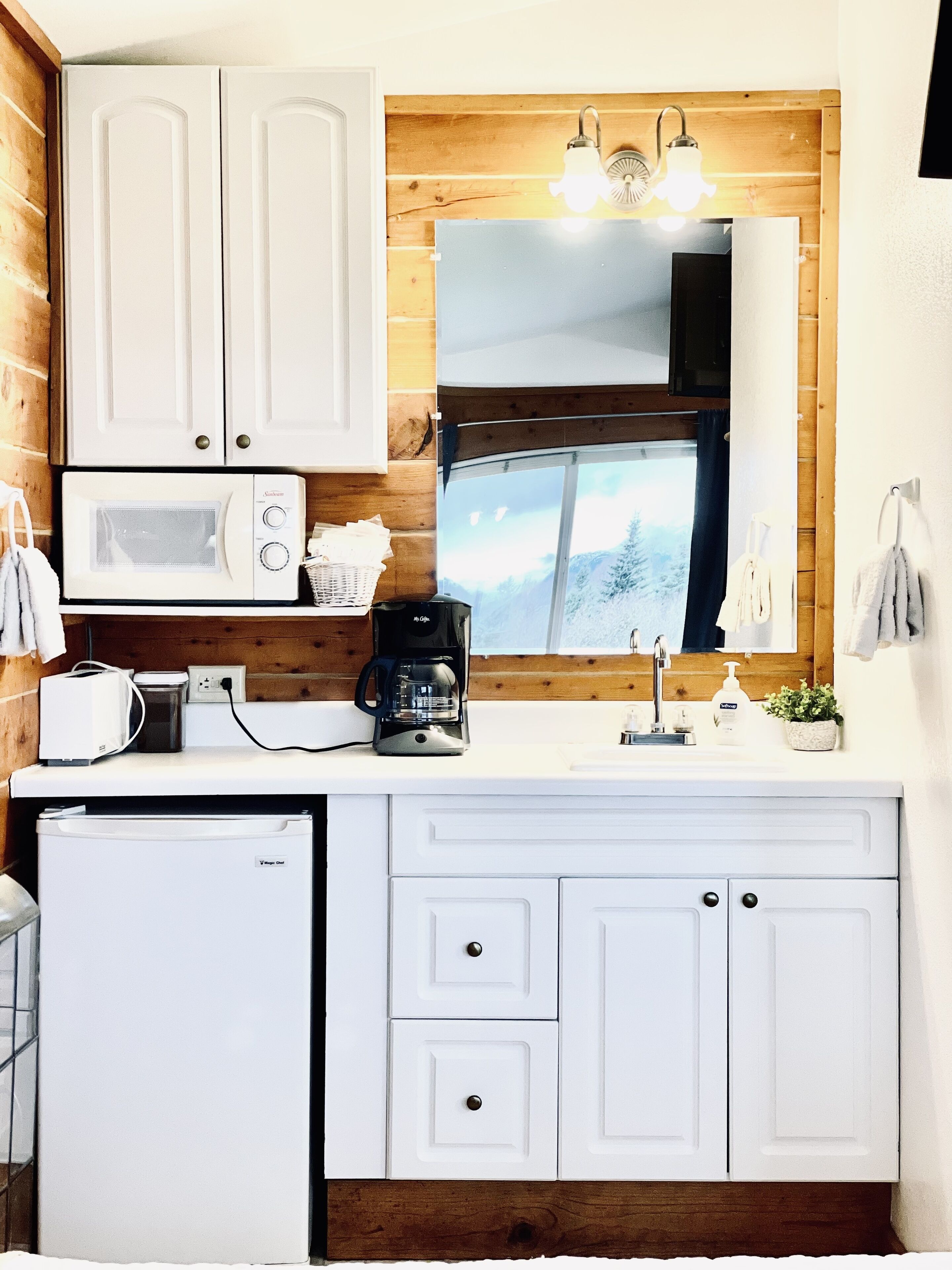 Una cocina compacta con gabinetes blancos, microondas, cafetera y refrigerador pequeño.