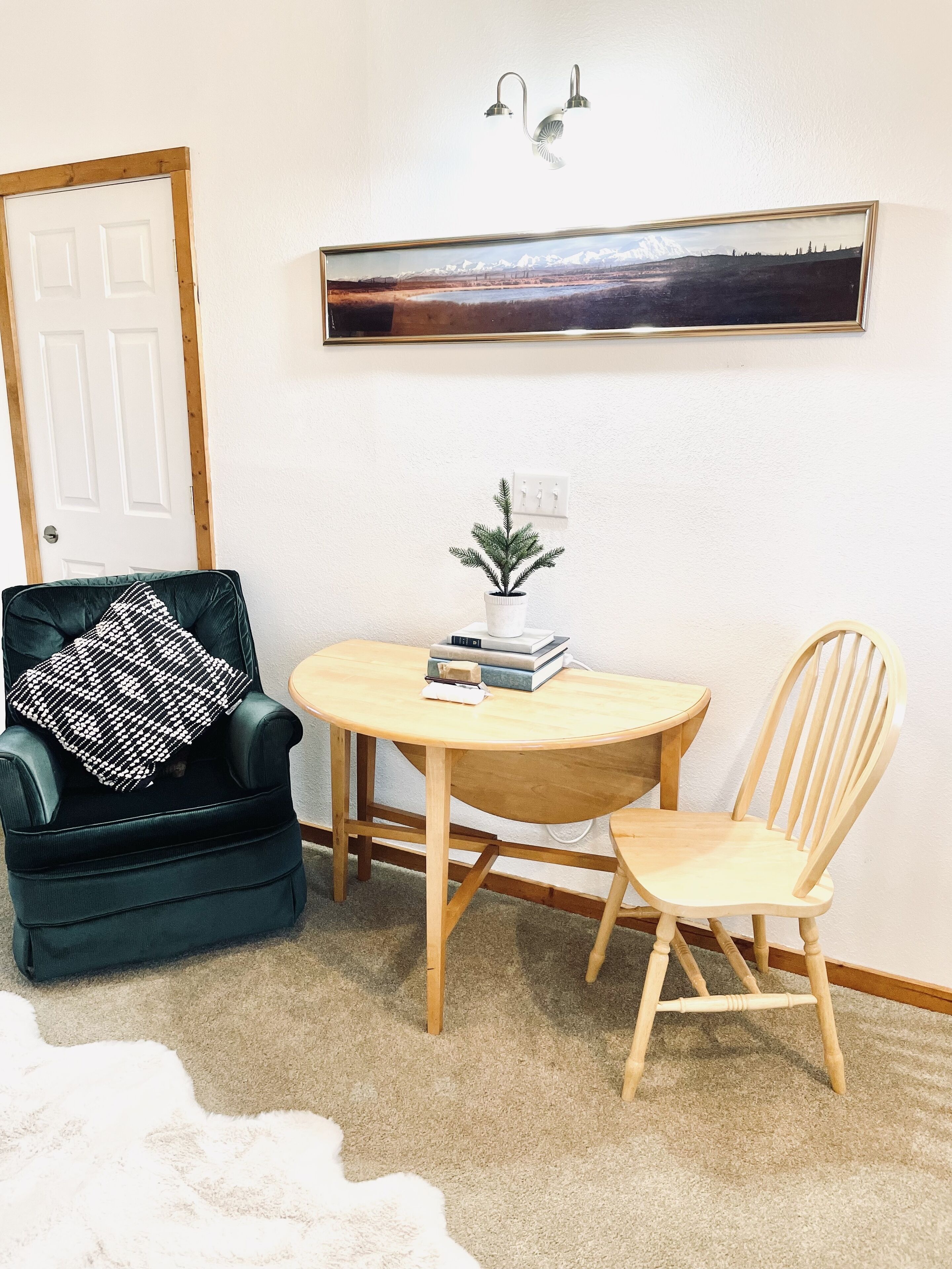 Een kamer met een donkergroene fauteuil, een witte stoel, een klein rond tafeltje met boeken en een plant, een lijst met een landschapsafbeelding, en een witte deur.