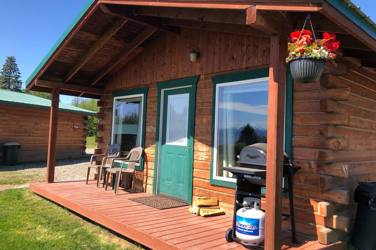 A wooden cabin with a porch, two chairs, a grill, and a propane tank.