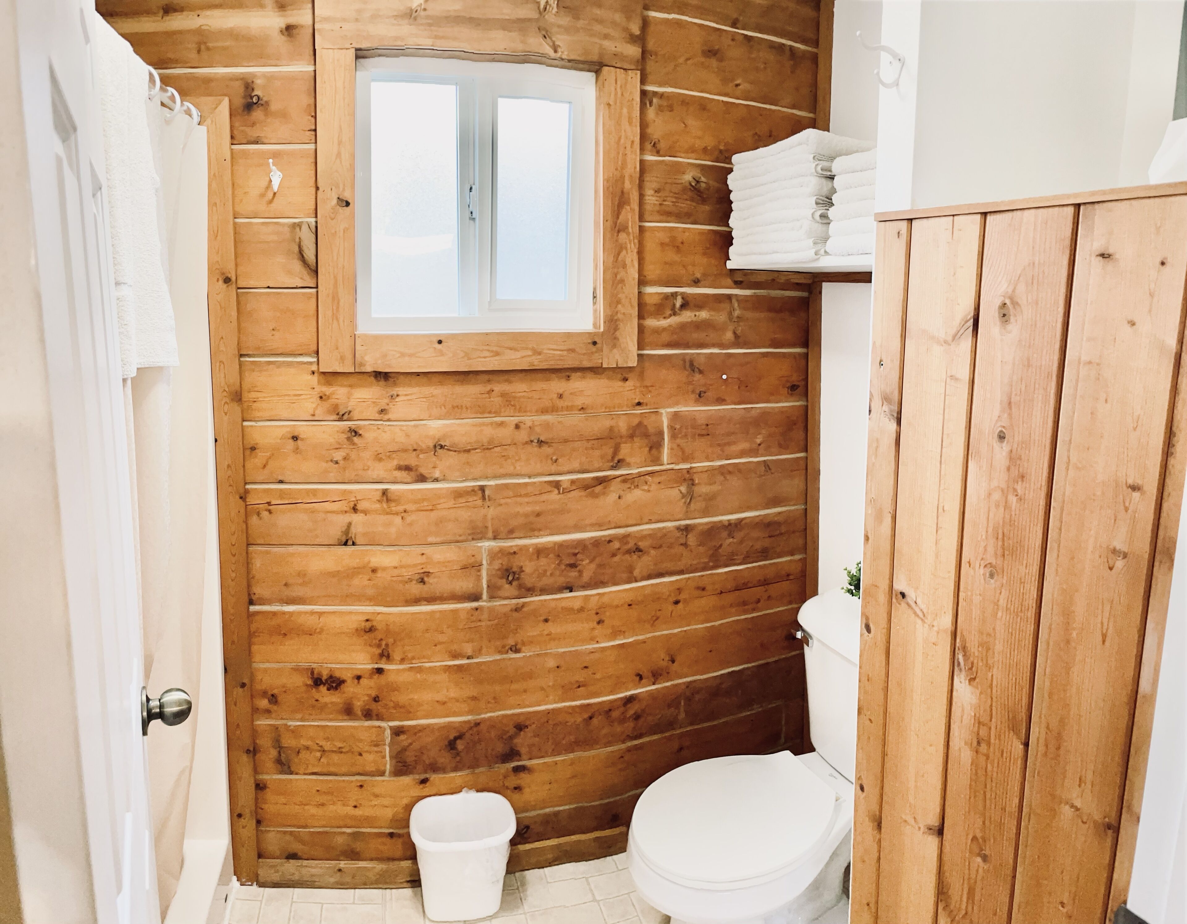 Une salle de bain avec des murs en bois, des toilettes blanches, une petite fenêtre et un seau blanc.