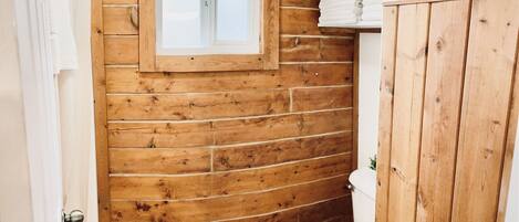 Une salle de bain avec des murs en bois, des toilettes blanches, une petite fenêtre et un seau blanc.