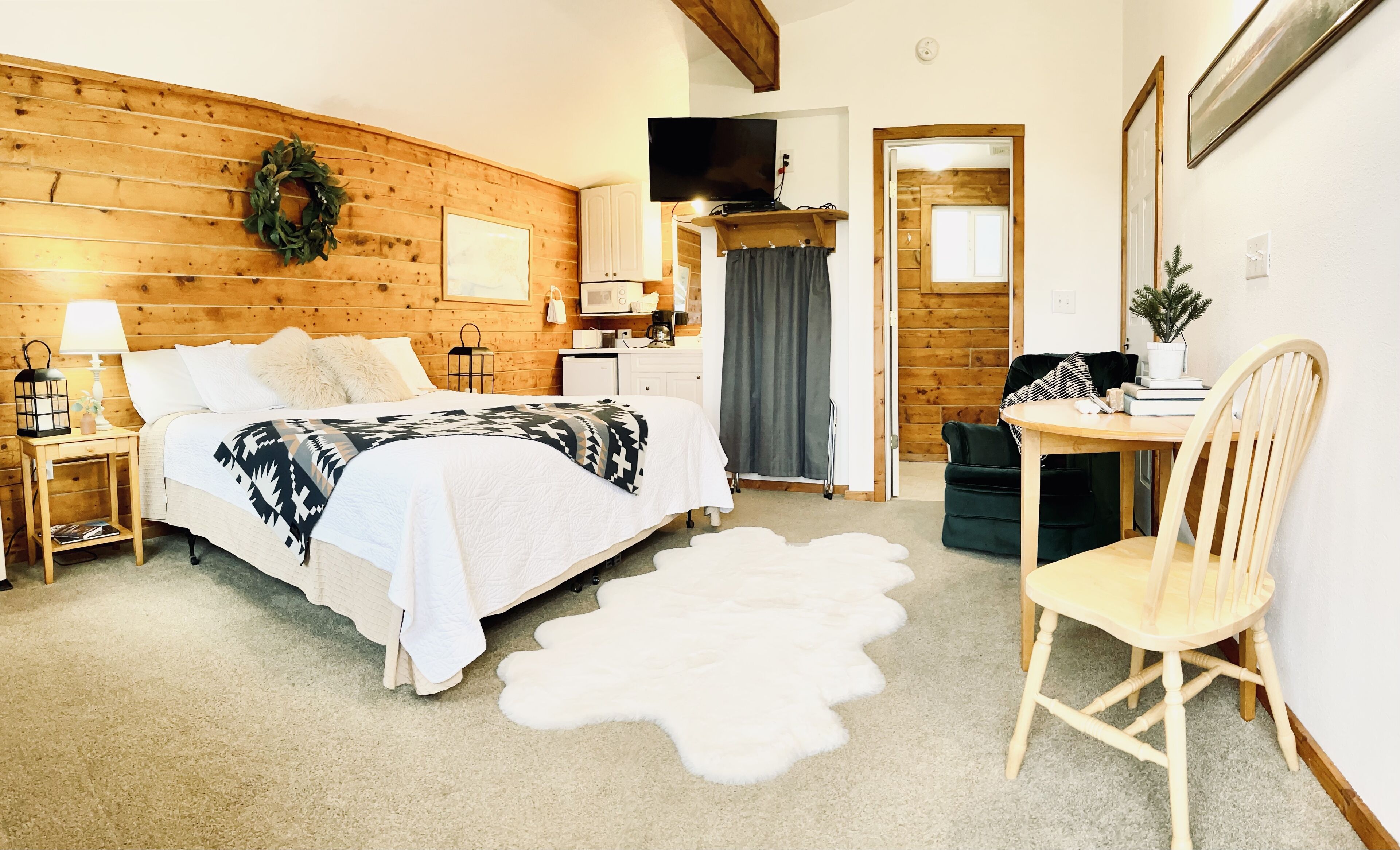 A cozy room with a wooden wall, a bed with a patterned blanket, a desk with a chair, and a television on the wall.