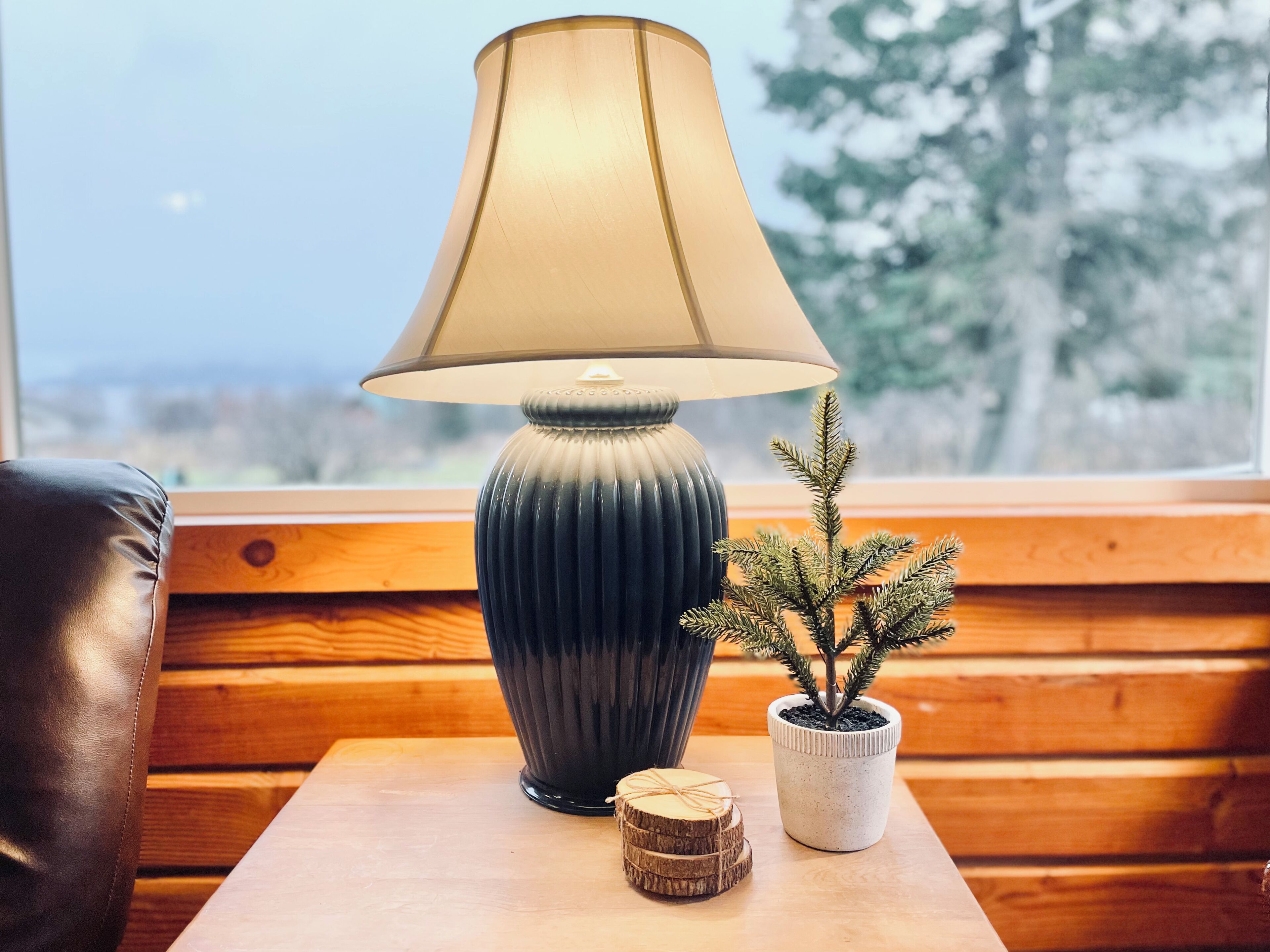 Eine Lampe mit geripptem Fuß und kegelförmigem Schirm, ein kleiner Blumentopf und zwei Holzuntersetzer auf einem Holztisch an einem Fenster.