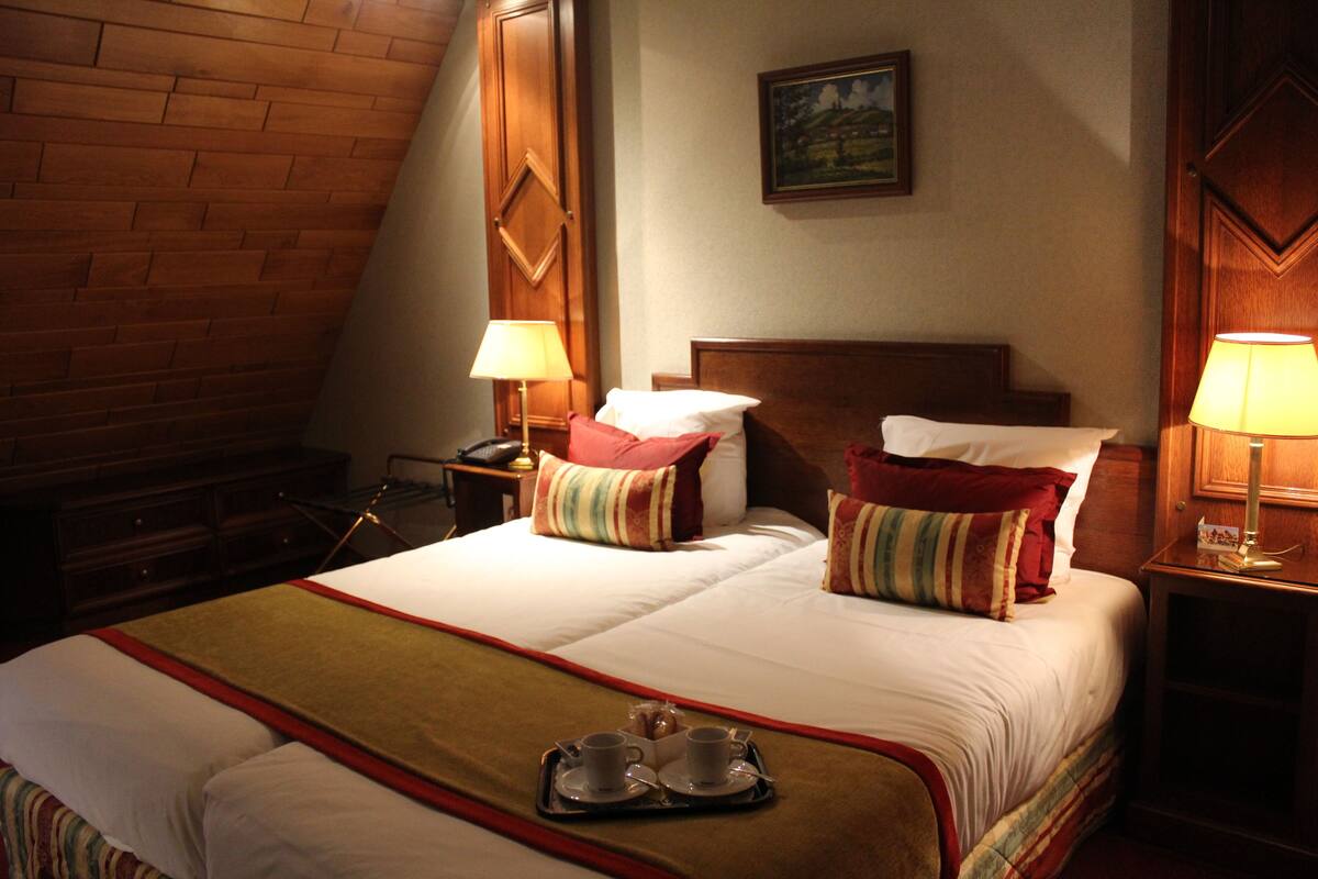 Habitación de hotel con una cama grande, dos mesitas de noche, un cabecero de madera, un cuadro en la pared y una bandeja con tazas y una tetera sobre la cama.