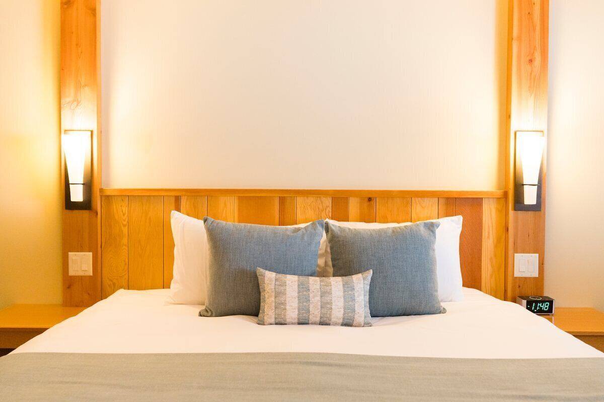 A neatly made bed with a white comforter and several pillows, including a striped one. There are two wall-mounted nightstands with a clock on one.
