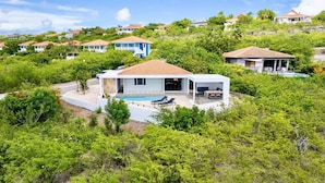 A modern house with a pool, surrounded by lush greenery and other houses in the background. - Coral Estate Rentals (St. Marie)