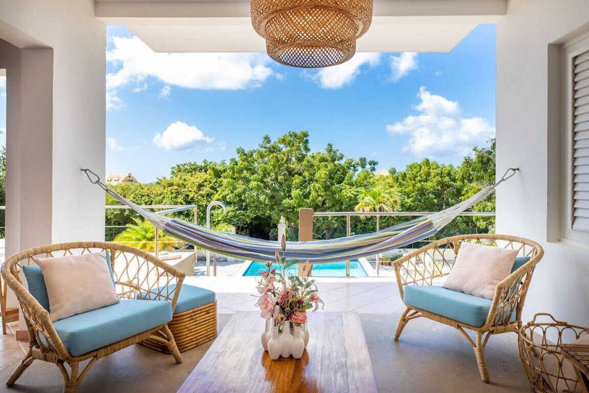 Ein Balkon mit Korbmöbeln, einer Hängematte und Blick auf einen Swimmingpool und Grünflächen.