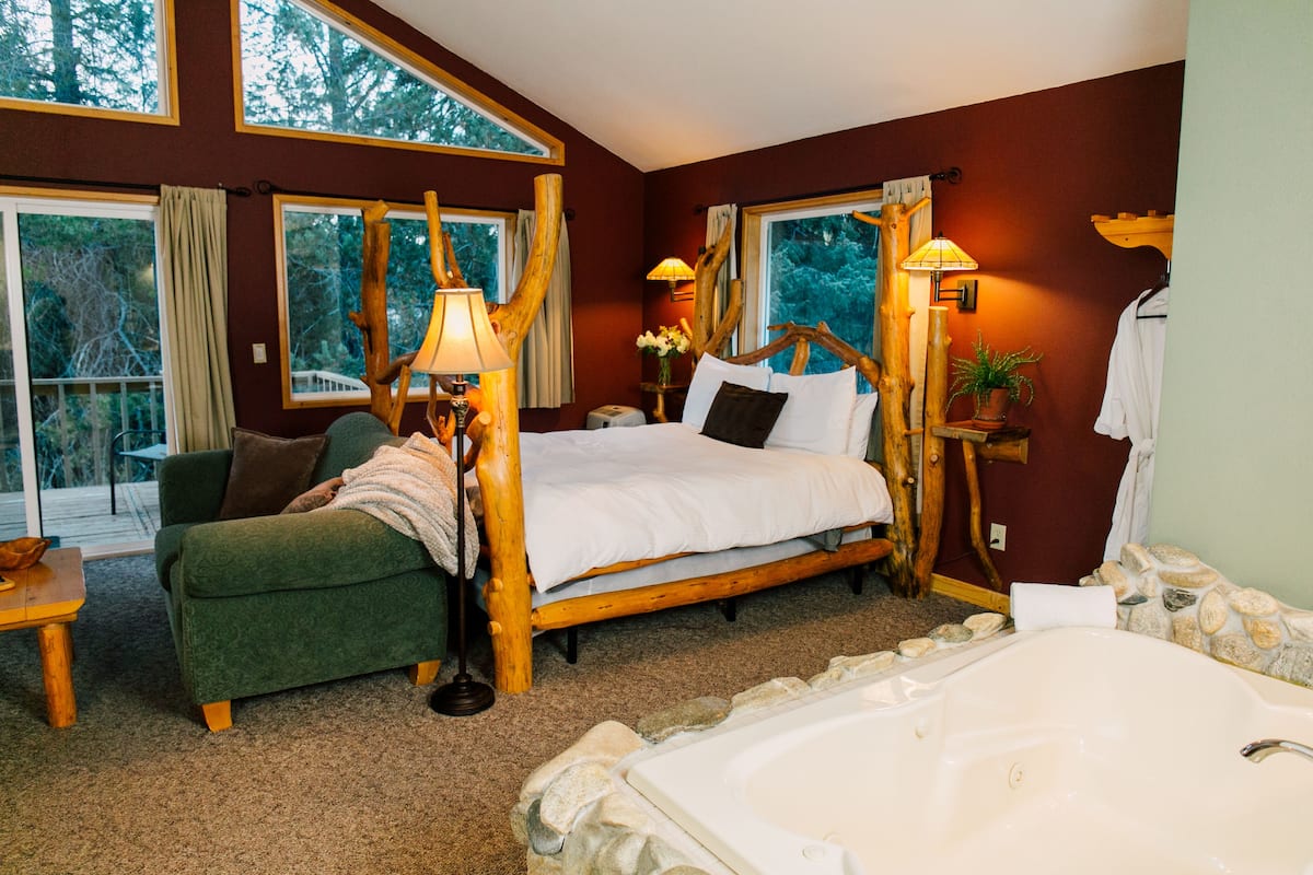 A bedroom with a large bed, a green armchair, a wooden headboard, a stone bathtub, and large windows with curtains.