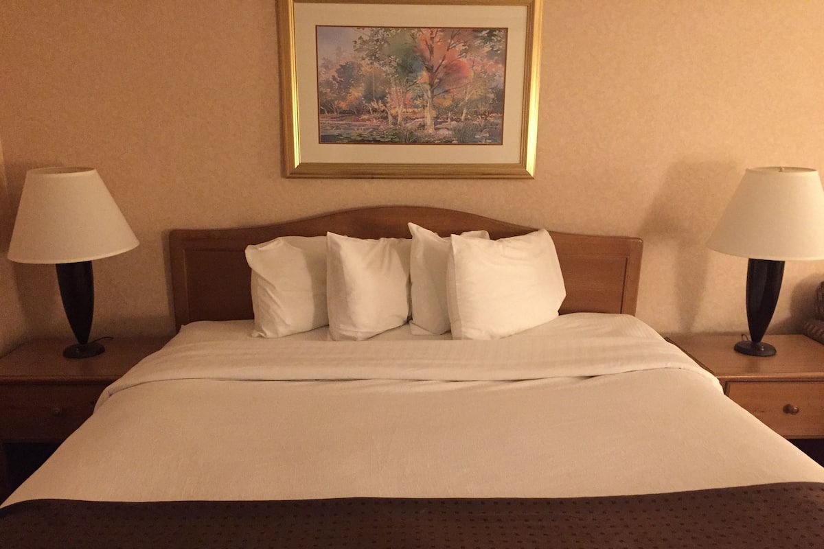 A hotel room with a bed, two bedside lamps, a framed picture on the wall, and a wooden headboard.