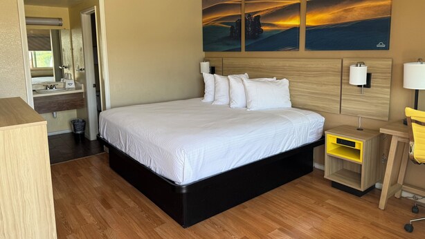 A hotel room with a large bed, a bedside table with a lamp, a mirror, a painting on the wall, and a wooden floor. - Days Inn by Wyndham Victorville/Hesperia I-15 & Palmdale Rd (Victorville)