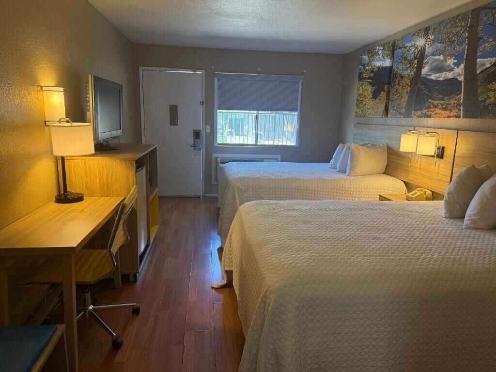 A hotel room with two beds, a desk with a lamp, a television, and a window with a view of trees.