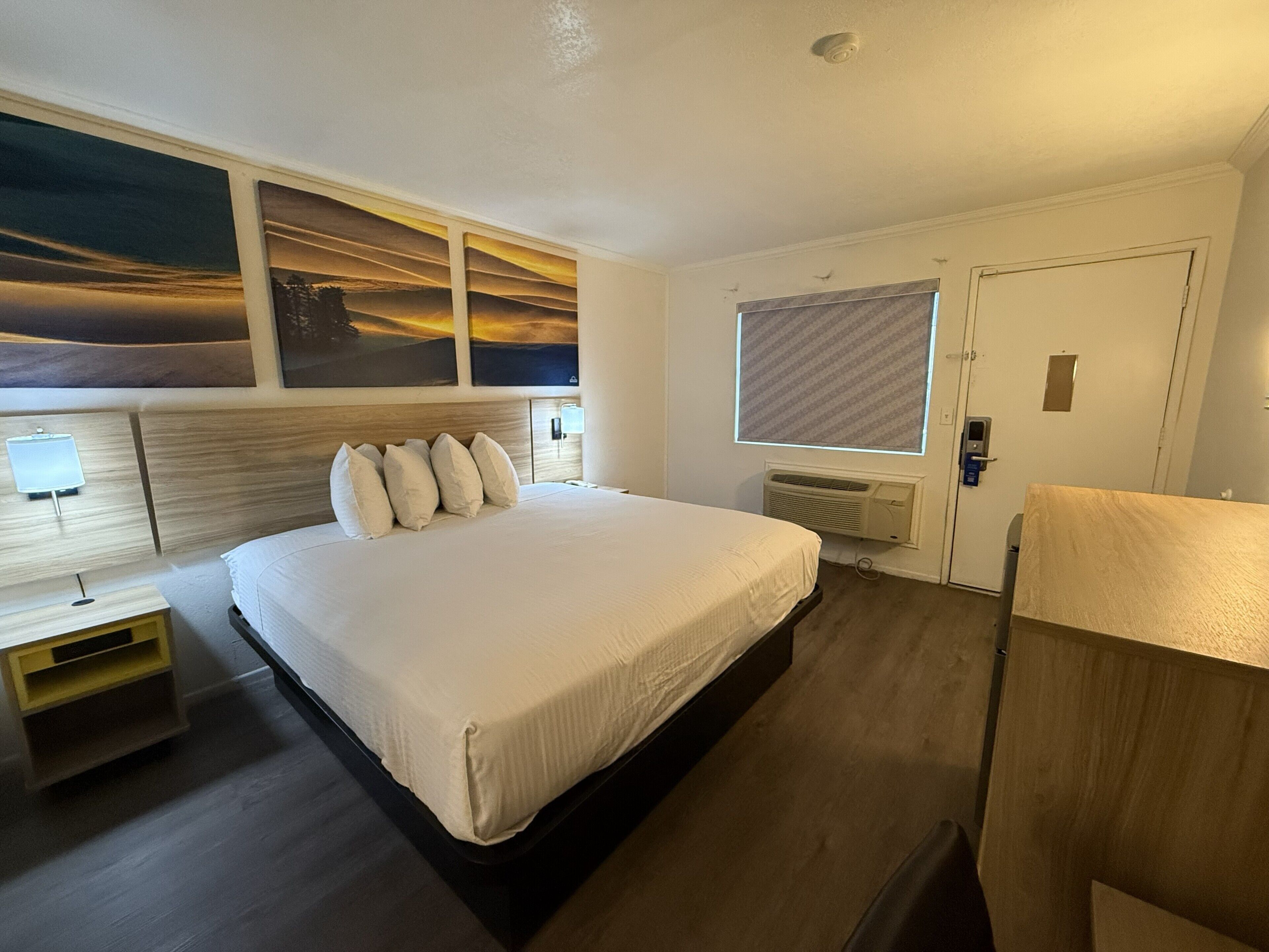 A hotel room with a large bed, a nightstand, a desk, a window with blinds, and a door.