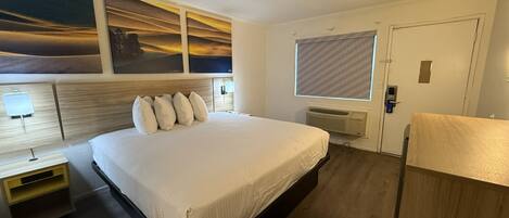 A hotel room with a large bed, a nightstand, a desk, a window with blinds, and a door.
