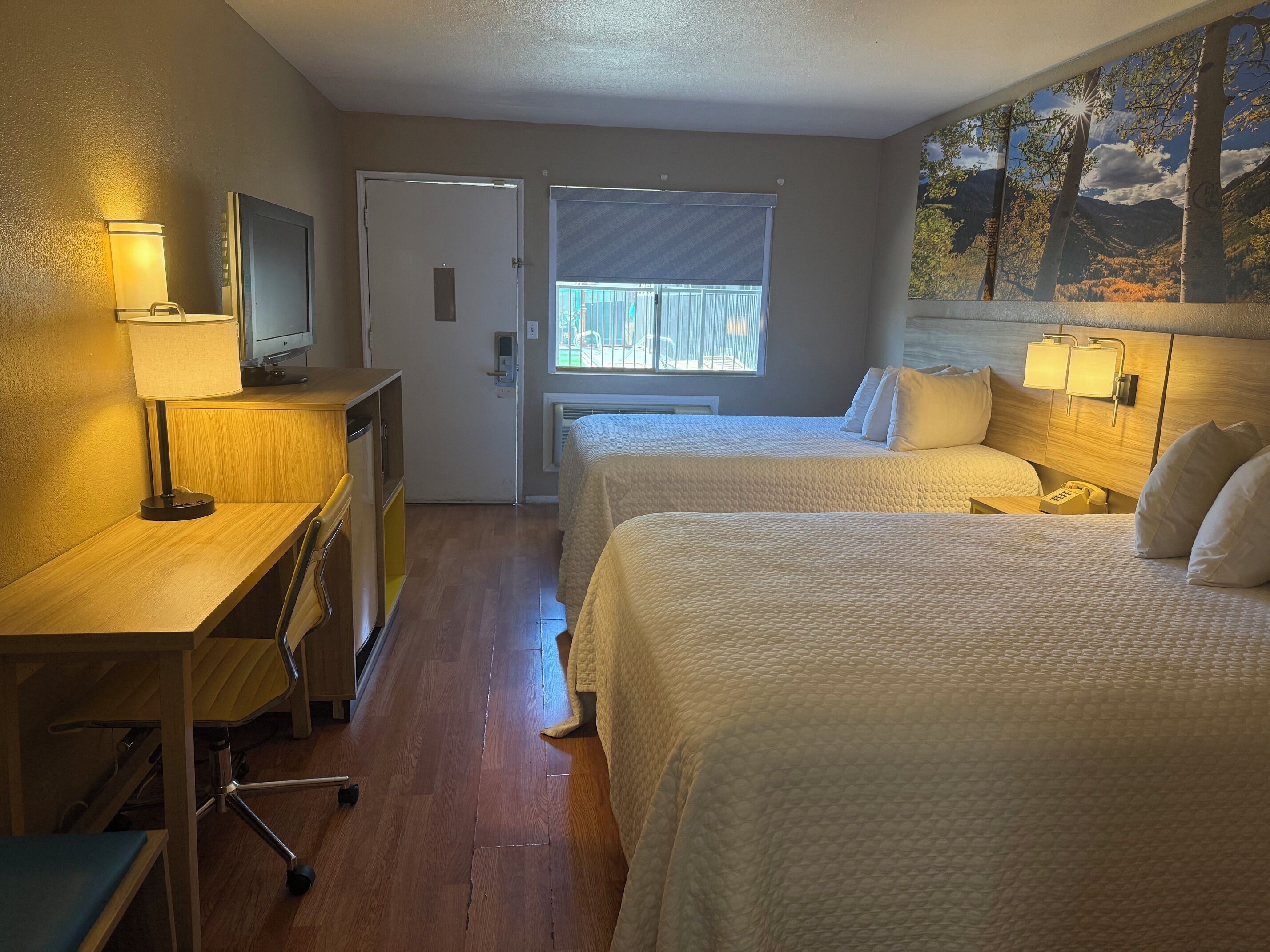 A hotel room with two beds, a desk with a lamp, a TV, a painting on the wall, and a window with blinds.