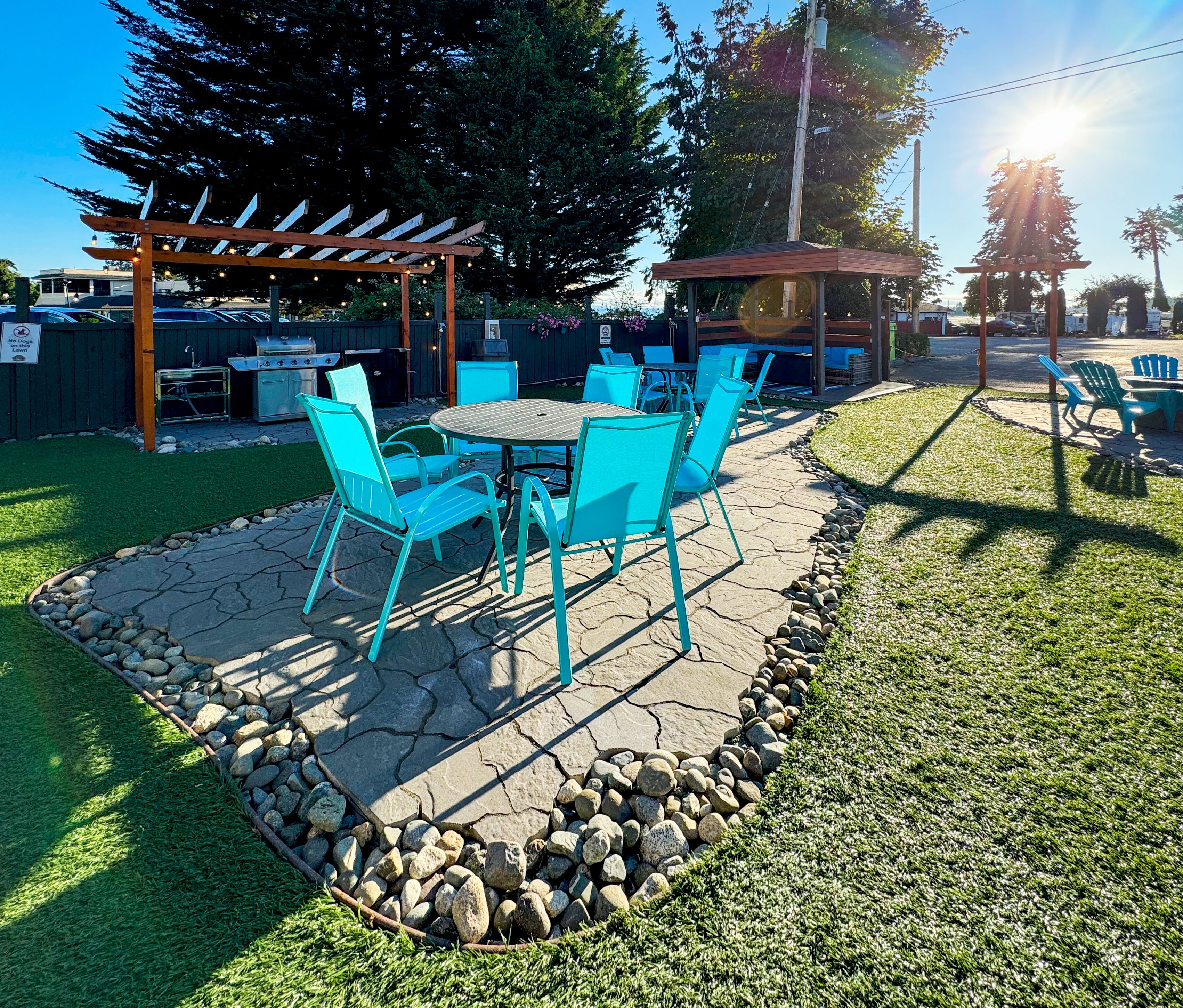 A patio area with a stone table and turquoise chairs, surrounded by a stone border and a grassy lawn.