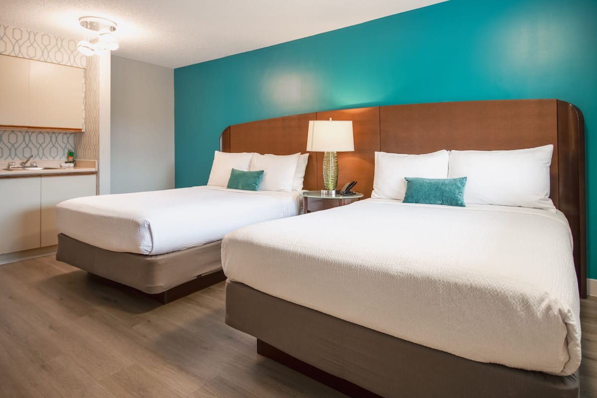 A hotel room with two beds, a nightstand with a lamp, a small table with a phone, and a bathroom with a mirror and towel rack.