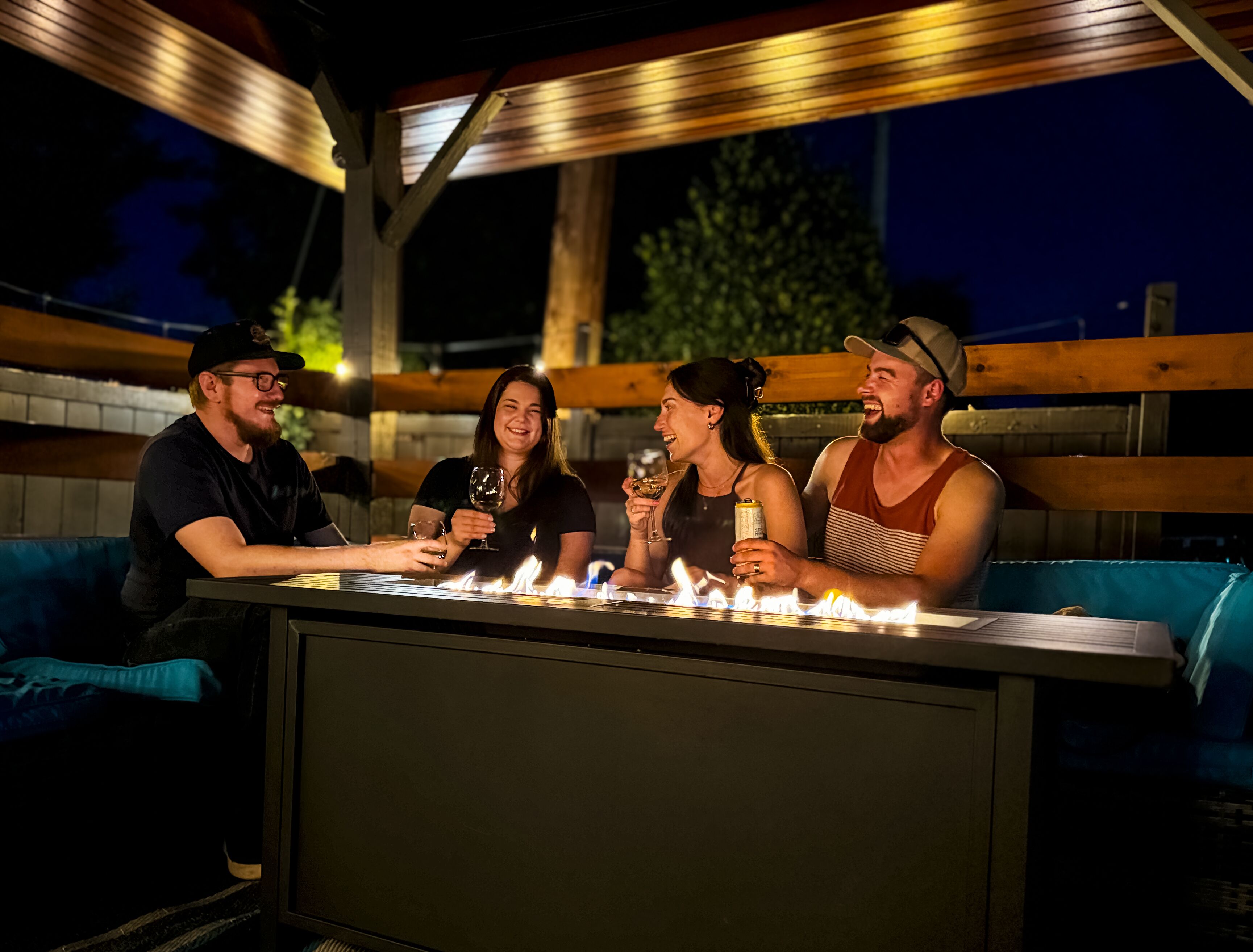 Un groupe de personnes est assis à un bar en plein air équipé d’un barbecue intégré, sous une pergola en bois, la nuit.