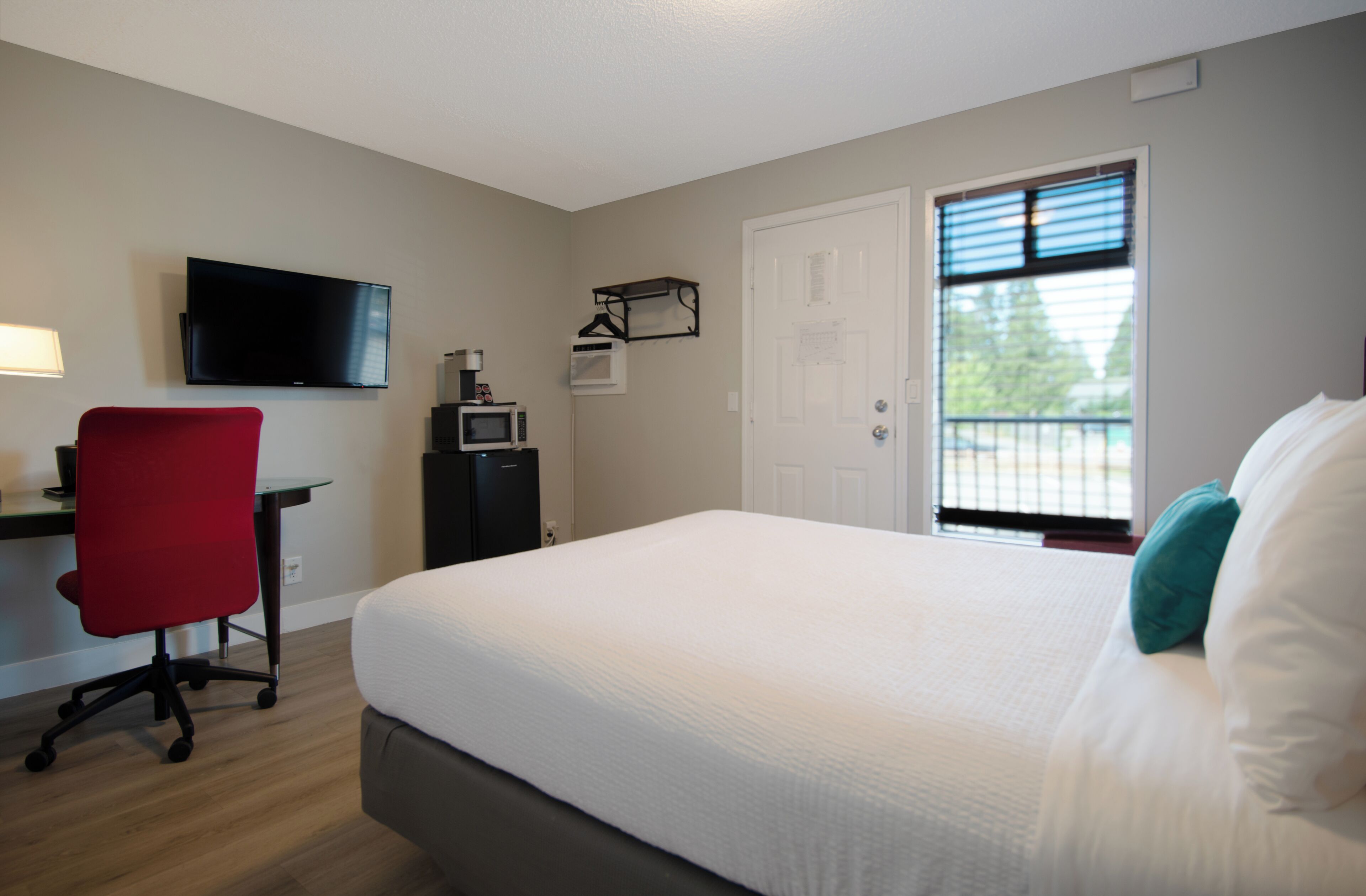 A bedroom with a bed, a desk, a chair, a TV, a door, and a window.