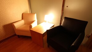 A room with a wooden floor, a beige armchair, a black armchair, and a small wooden table with a lamp. - Prince Hotel (Seoul)