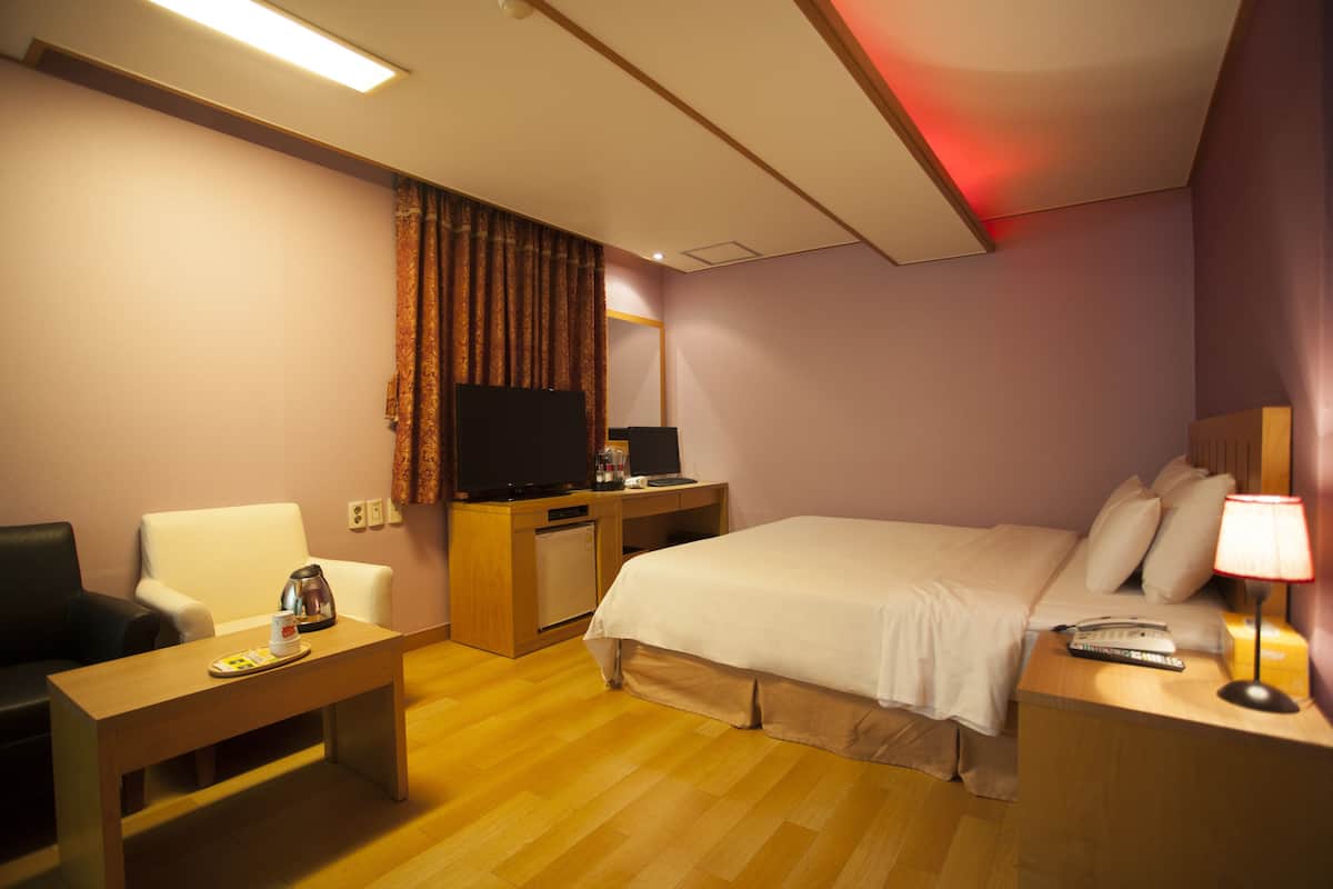 A hotel room with a bed, a desk, a chair, a television, and a nightstand with a lamp.