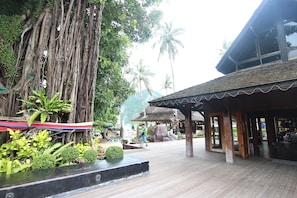A wooden deck with a covered seating area, surrounded by lush greenery and palm trees.