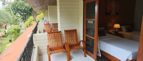 A balcony with wooden chairs, a bed with white bedding, and a view of greenery.