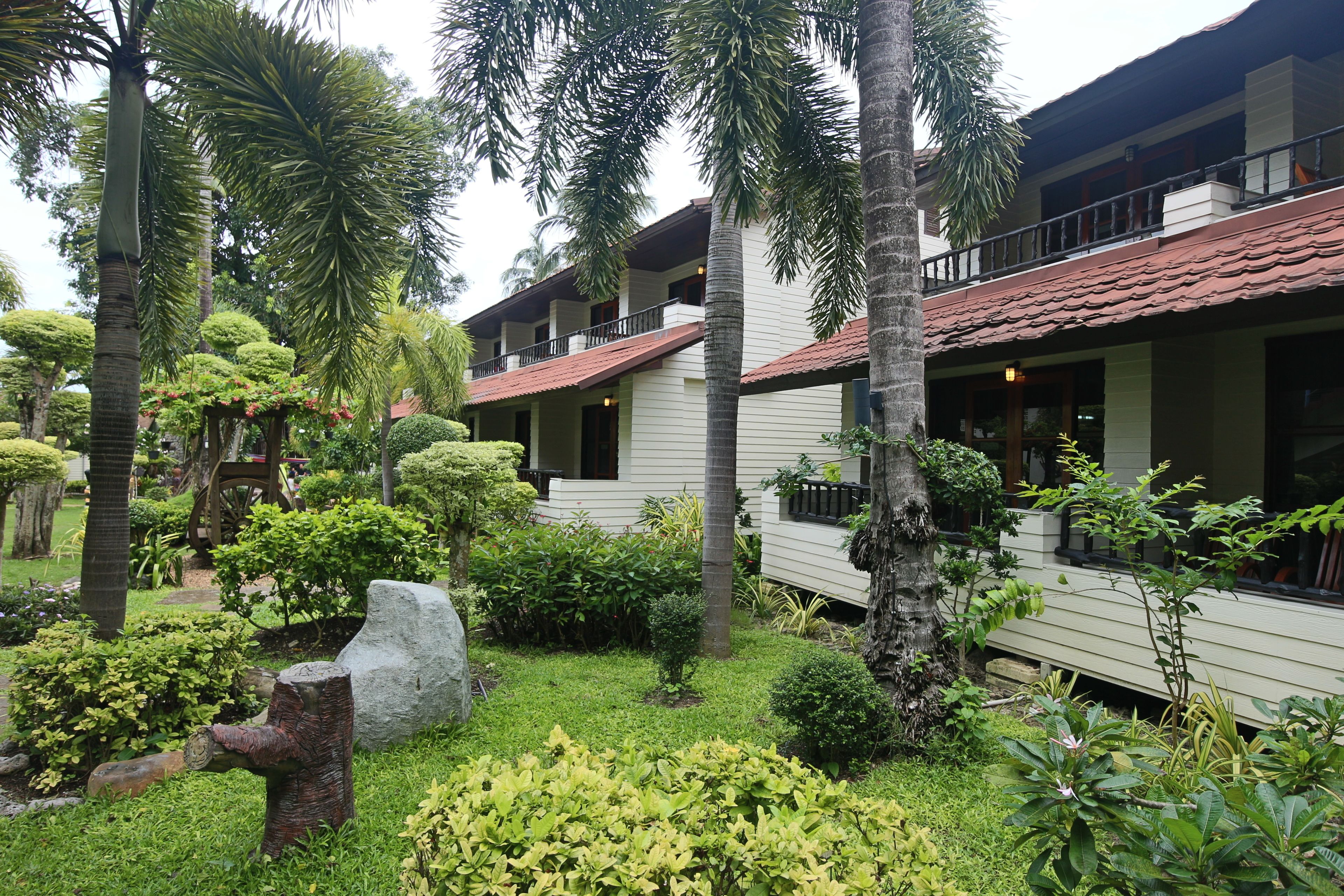 A hotel with a white exterior, red-tiled roof, and multiple balconies. The surrounding area includes well-maintained tropical landscaping with various trees and shrubs.