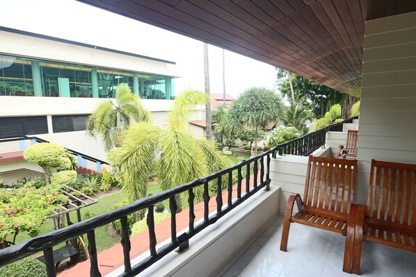 A balcony with a wooden railing, a bench, and a view of a garden with various plants and a building in the background.