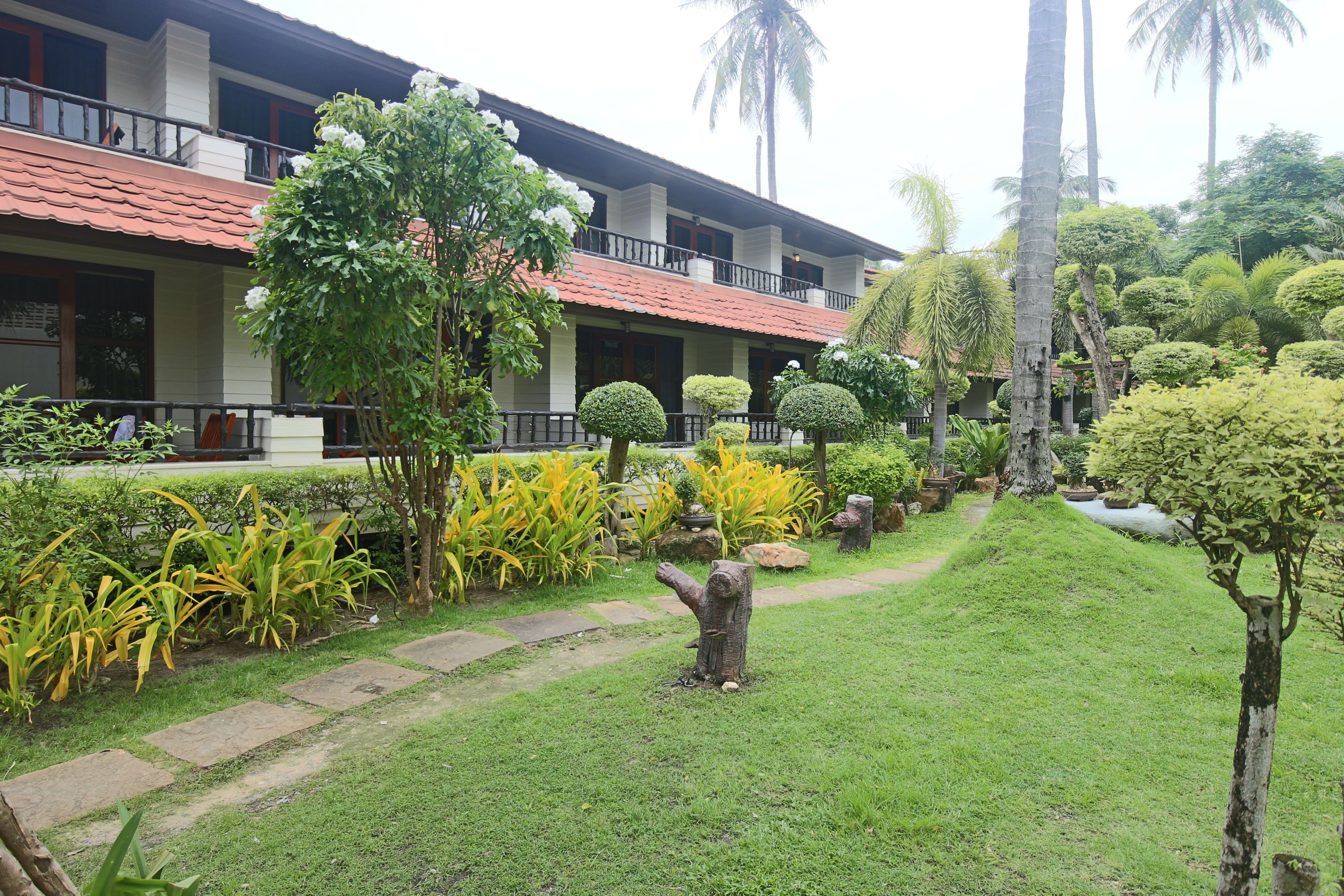 A hotel with a red-tiled roof, a balcony, and a well-maintained garden with various plants and a small pond.