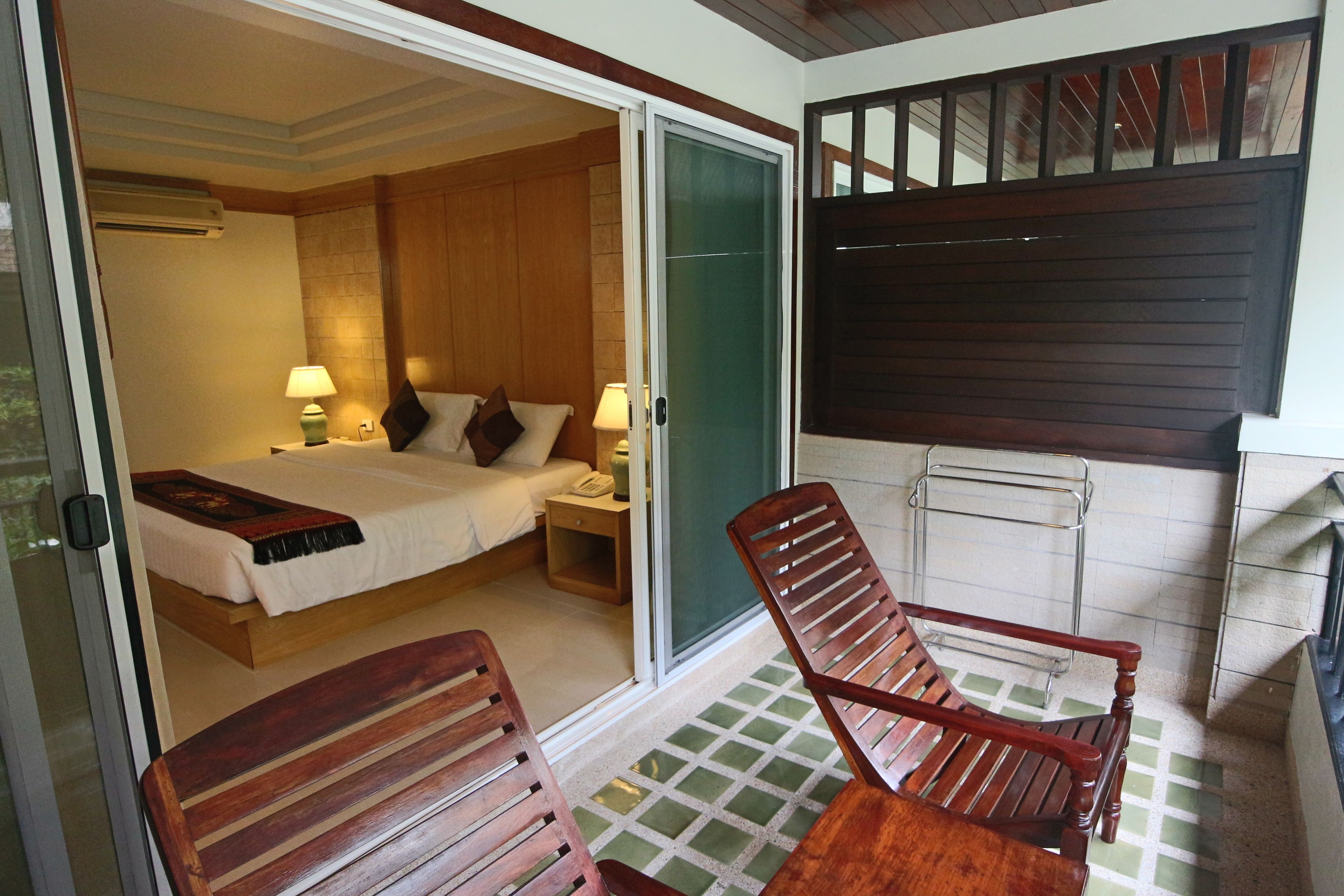 Ein Schlafzimmer mit einem Bett, Nachttischen und einer Schiebetür zu einem Balkon mit Holzstühlen und Blick ins Freie.