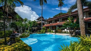 Une piscine entourée d’une végétation luxuriante et de palmiers, avec un bâtiment doté d’un toit rouge en arrière-plan.