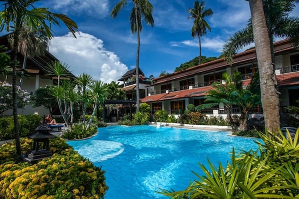 A swimming pool surrounded by lush greenery and palm trees, with a building featuring a red roof in the background.