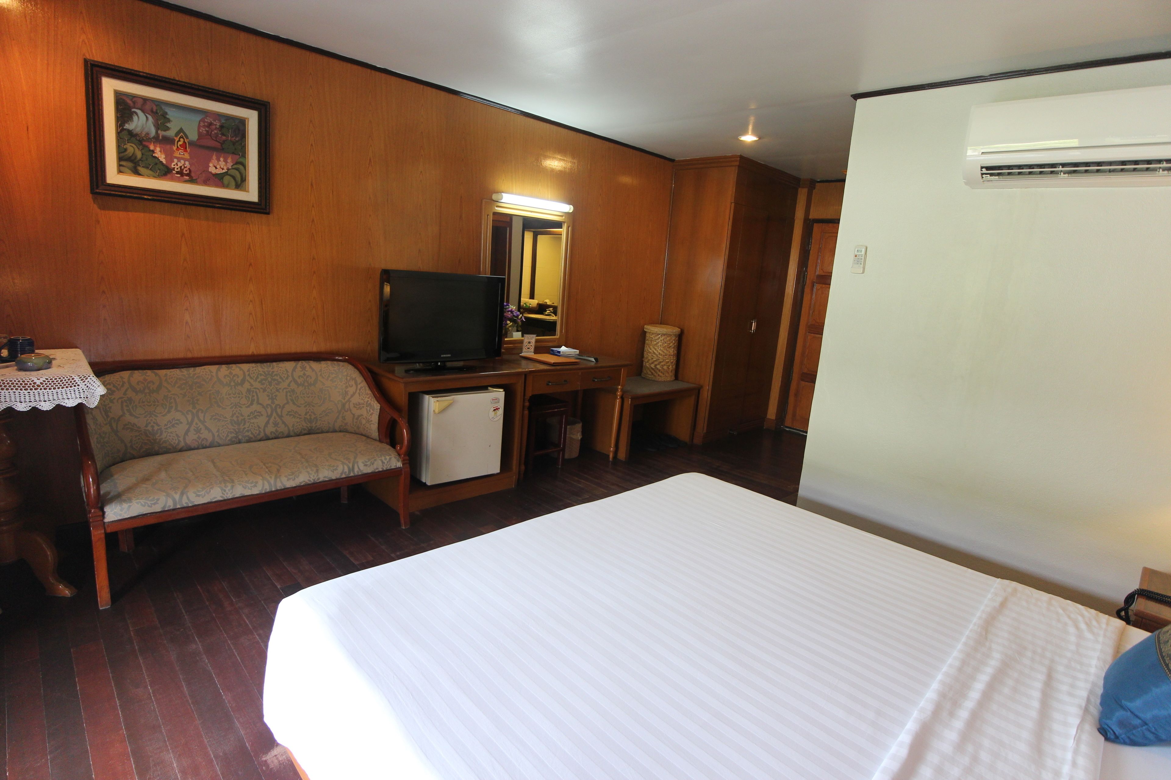 A hotel room with a bed, a sofa, a desk with a television, a mirror, and an air conditioning unit.