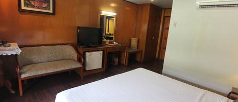 A hotel room with a bed, a sofa, a desk with a television, a mirror, and an air conditioning unit.