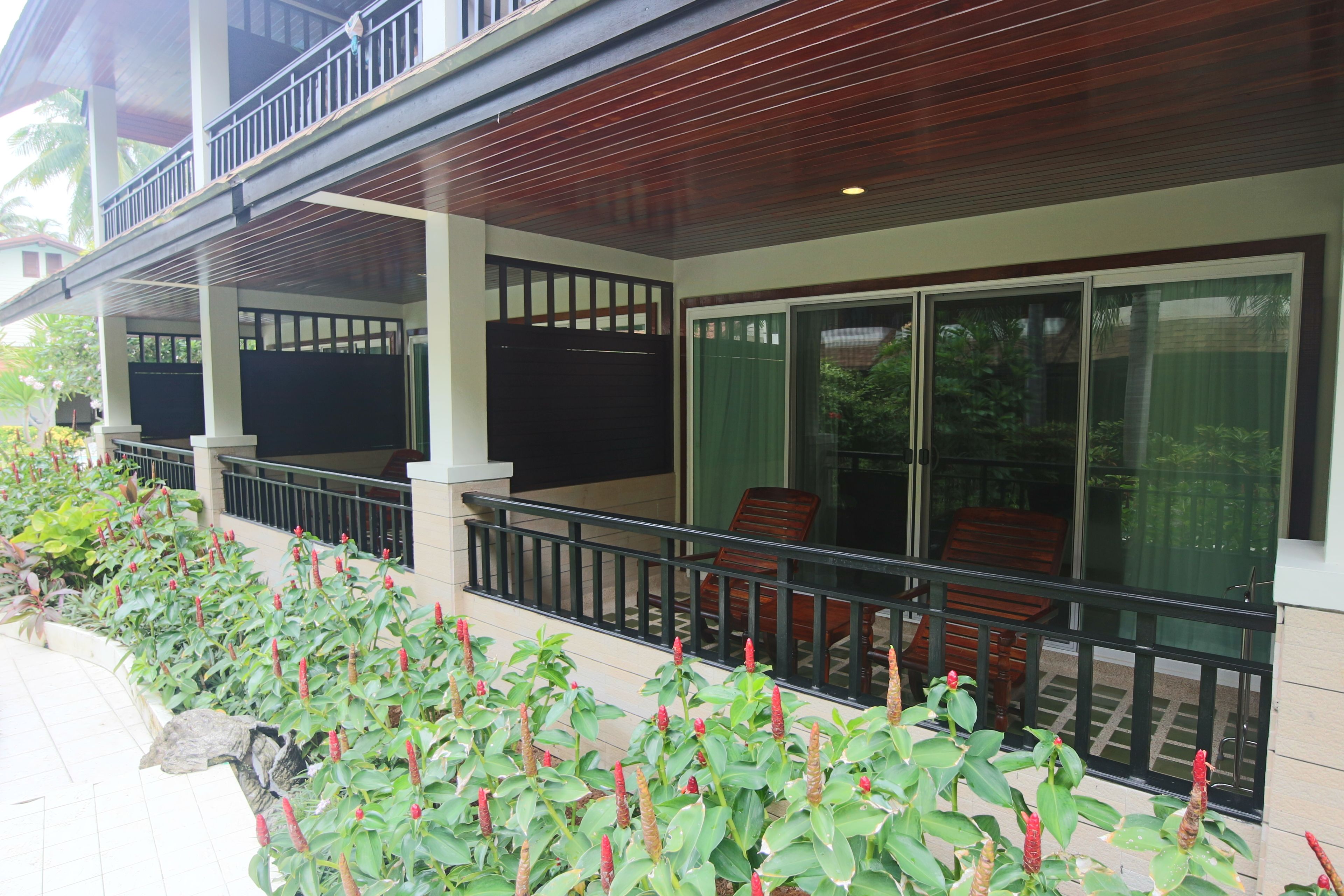 A covered outdoor area with a wooden ceiling, a balcony with chairs, and a row of plants with red flowers.