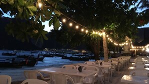 Un restaurant situé en bord de mer, avec une terrasse extérieure illuminée de guirlandes lumineuses, offrant une vue sur l’océan et des bateaux à l’horizon.