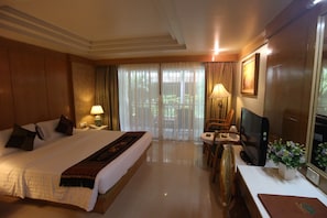 A hotel room with a large bed, a desk with a television, a chair, a table, and a window with curtains.