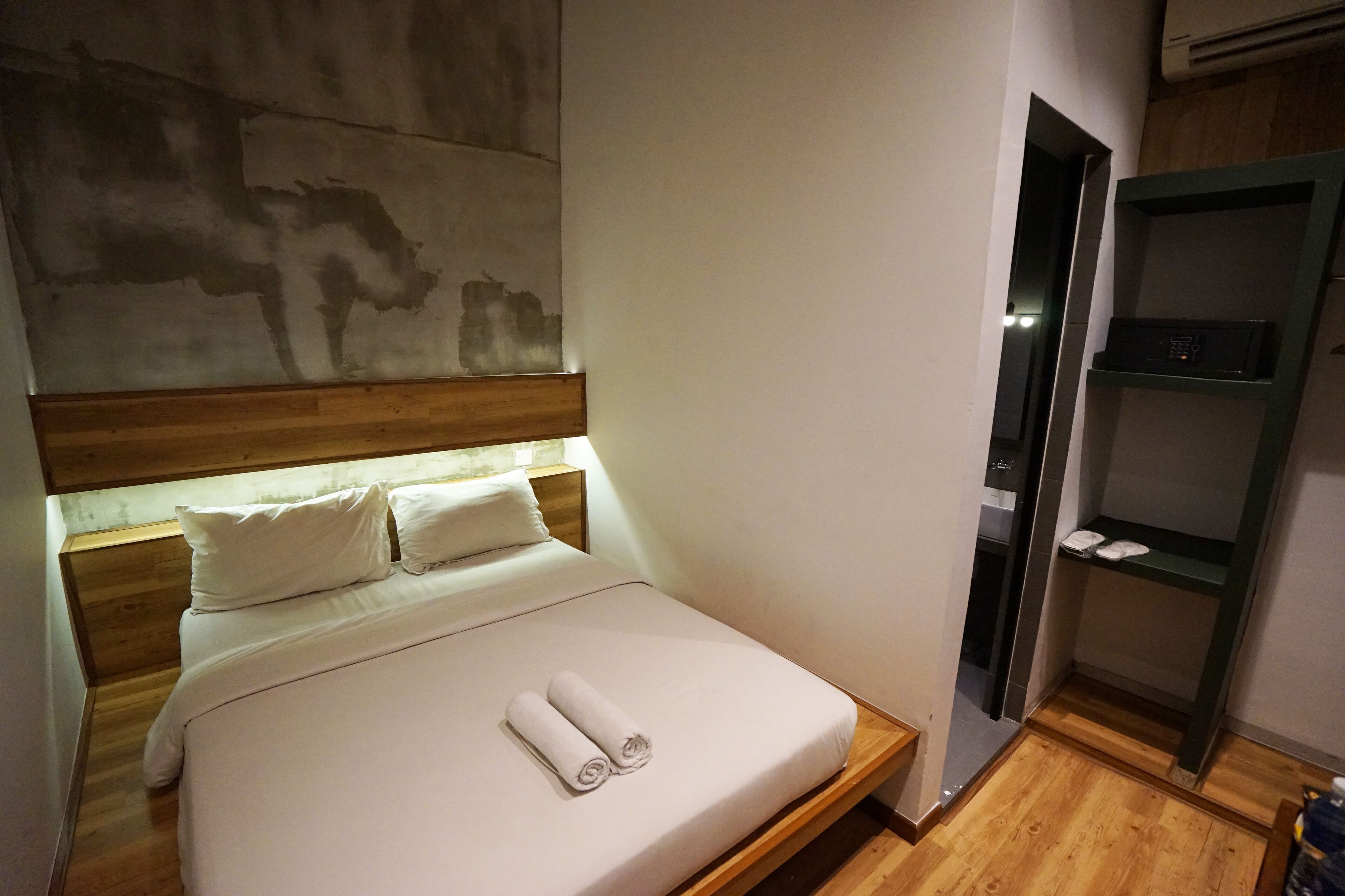 Una cama bien hecha con sábanas blancas y una toalla enrollada, un cabecero de madera y una estantería de pared con un objeto pequeño.