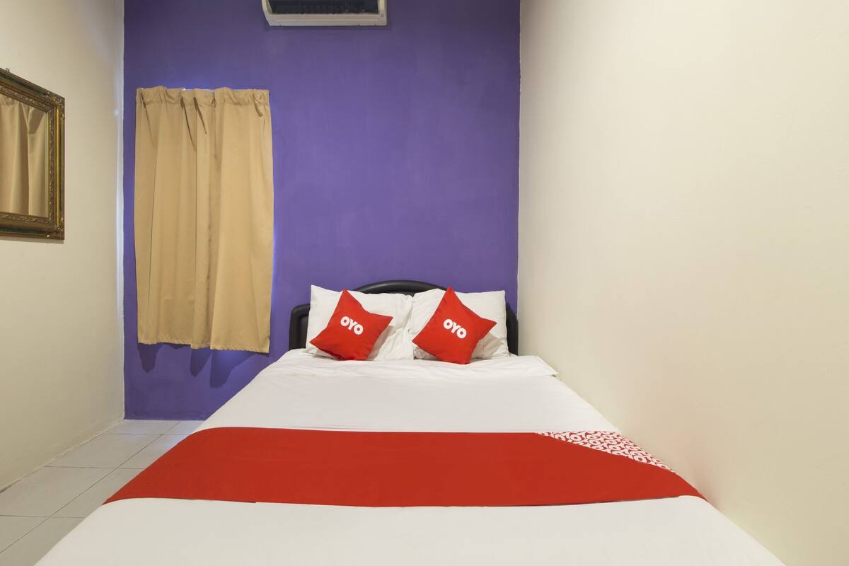 A hotel room with a bed, a window with beige curtains, and a purple wall.