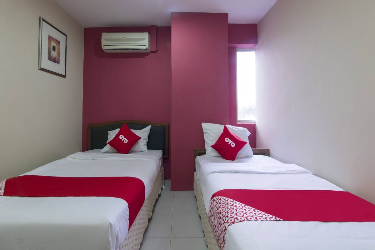 A hotel room with two beds, each with a red blanket and pillows, a window, and an air conditioning unit on the wall.