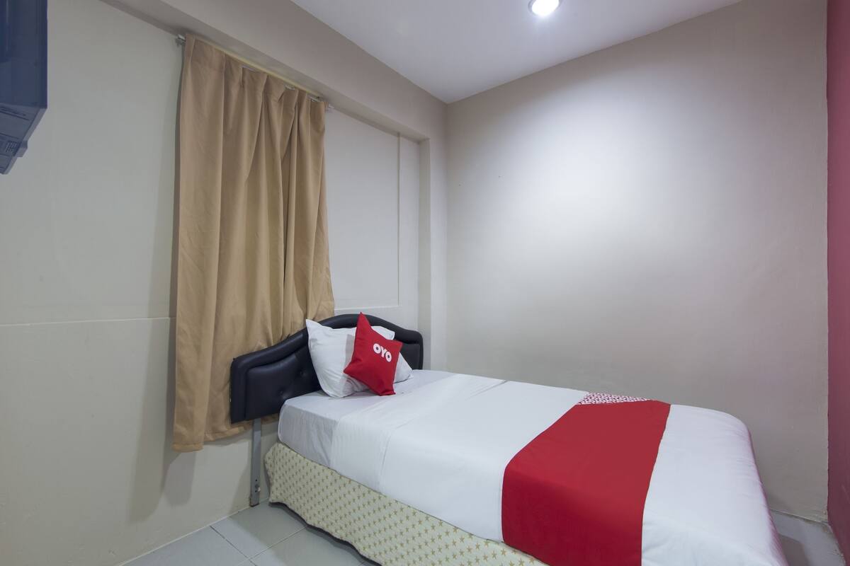 A hotel room with a bed, beige curtains, a red and white bedspread, and a white wall.