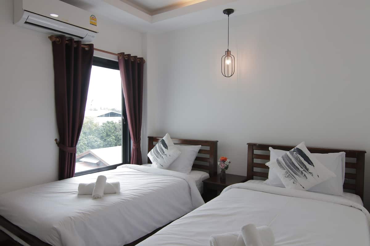 A hotel room with two beds, white bedding, a window with curtains, an air conditioning unit, a bedside table with a vase of flowers, and a hanging light fixture.