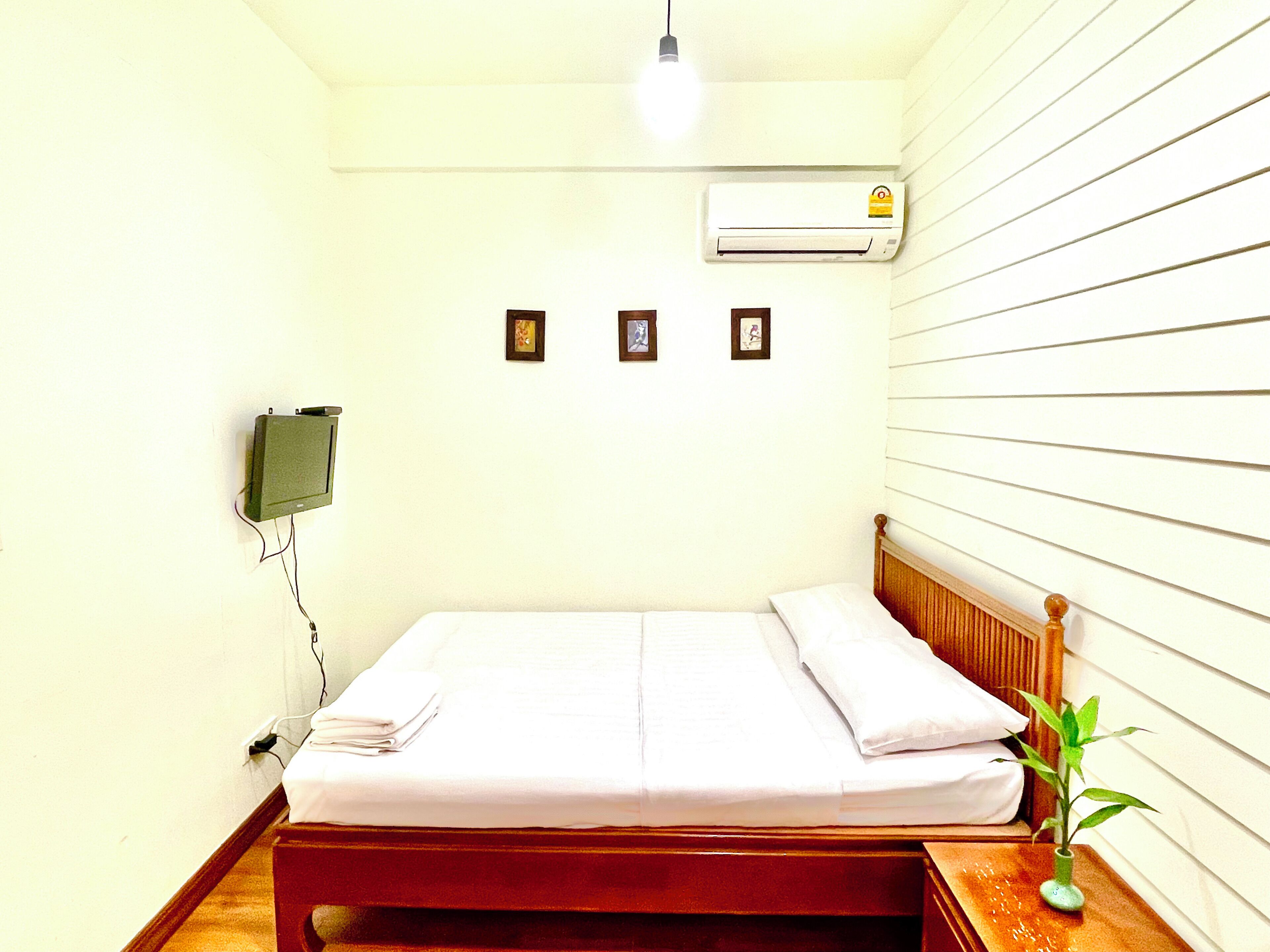 Un lit simple avec une tête de lit en bois, un climatiseur fixé au mur, une petite télévision accrochée au mur et une plante en pot sur la table de chevet.